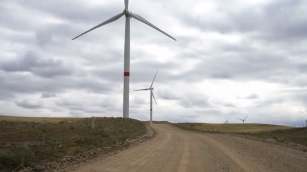 Tarladaki büyük bir rüzgar türbininin kanatlarını tarla yolunun yakınındaki bulutlu mavi gökyüzünün arka planına doğru hareket ettir. Alternatif enerji kaynakları. Rüzgarlı park. Ekolojik enerji. — Stok video