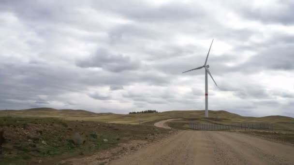 Ruch łopaty dużej turbiny wiatrowej w polu na tle zachmurzonego niebieskiego nieba w pobliżu drogi polowej. Alternatywne źródła energii. Park wiatrów. Energia ekologiczna. — Wideo stockowe
