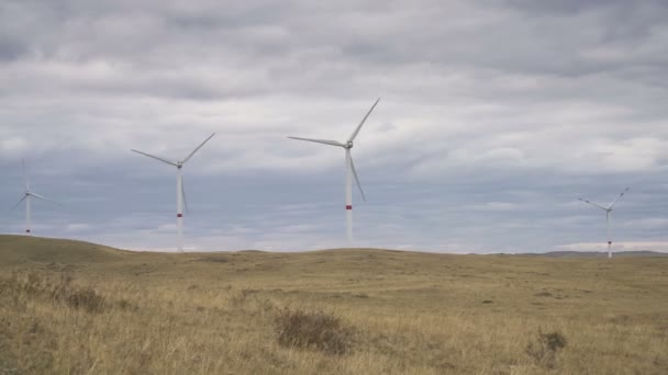 Ufukta bulutlu gri gökyüzü ve güzel tepeleri olan bir tarladaki büyük bir rüzgar türbininin kanatlarını hareket ettir. Alternatif enerji kaynakları. Rüzgarlı park. Ekolojik enerji. Sanayi. — Stok video