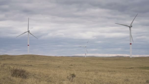 Bewegen Sie die Flügel einer großen Windkraftanlage auf einem Feld vor einem wolkenverhangenen grauen Himmel am Horizont mit einem wunderschönen Hügel. Alternative Energiequellen. Windiger Park. Ökologische Energie.Industrie — Stockvideo