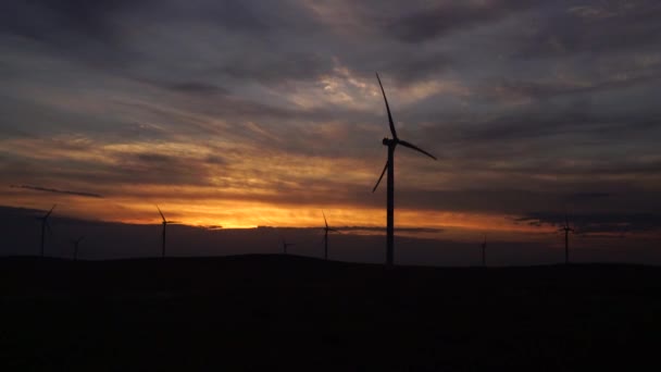 Bewegen Sie die Flügel einer großen Windkraftanlage auf einem Feld vor dem Hintergrund eines orangefarbenen Sonnenuntergangs am Horizont mit einem wunderschönen Hügel. Alternative Energiequellen. Windiger Park. Ökologische Energie.Industrie — Stockvideo