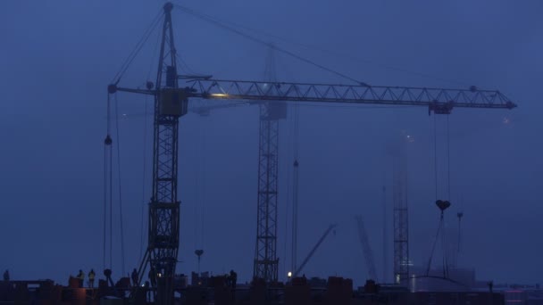 Construction site at dusk with purple sky. Silhouette of high tower cranes works on high-rise residential building site, lifts load. — Stock Video