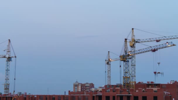 Construction site at dusk with purple sky. Silhouette of high tower cranes works on high-rise residential building site, lifts load. — Stock Video