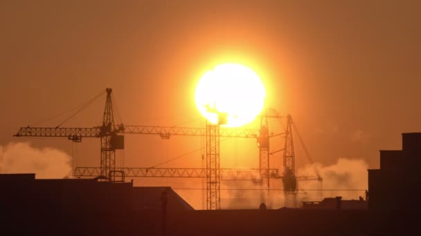 Construction site at orange sunset. Silhouette of high tower cranes works on high-rise residential building site, lifts load. — Stock Video