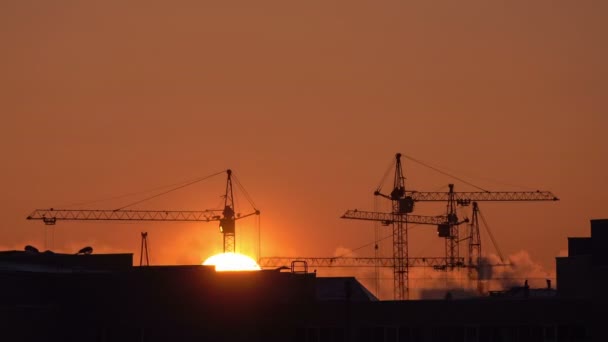 Site de construction au coucher du soleil orange. Silhouette de grues à tour hautes travaux sur le site de construction résidentielle de grande hauteur, charge ascenseurs. — Video
