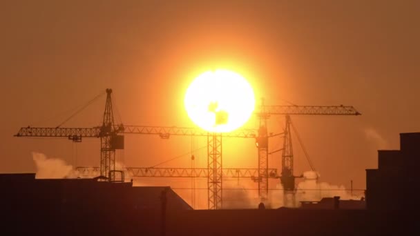 Construction site at orange sunset. Silhouette of high tower cranes works on high-rise residential building site, lifts load. — Stock Video