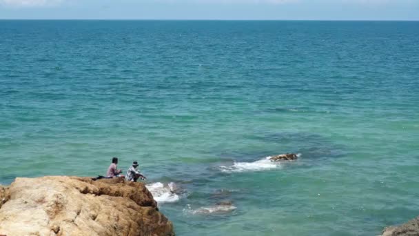 Zwei unbekannte Fischer sitzen auf einem braunen Felsen am Meer. — Stockvideo