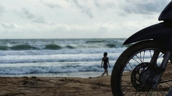 Silhouet van het voorwiel van motorfiets geparkeerd op strand voor Oceaan, golven spatten op de kust. Silhouet van niet-identificeerbare vrouw lopen op het strand. Rechtenvrije Stockfoto's