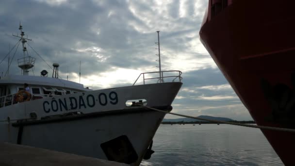 Nave blanca a Con Dao atada al amarradero por una cuerda gruesa. Arco de otro gran barco rojo visible frente al cielo nublado . — Vídeos de Stock