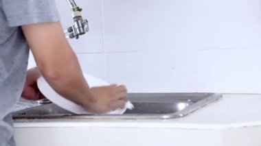 Asyalı genç adam ayakta duruyor ve mutfakta bulaşıkları yıkıyor.