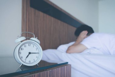 Yataktaki çalar saatin gürültüsünden kafasını ve kulaklarını yastıkla kapatan bir adam, sinirlenmiş..