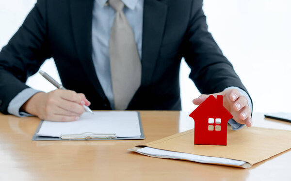 House on document envelope with man signing purchase documents in background. while hand complete the insurance policy, rental documents and loan document. concept guy buying new house