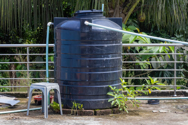 Большой резервуар для черной пластиковой воды, установленный на крыше в деревне Бангладеш. Танк окружен неопрятным мхом, сорняками и выброшенным бытовым мусором, нуждающимся в уборке..