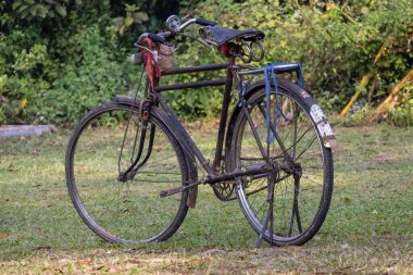 Klasik, iyi kullanılmış, pas ve yıpranmayı gösteren eski model bir bisiklet, yeşil bir çim tarlasına park edilmiş. Bangladeş / Güney Asya kırsalında yerel taşımacılık ve sürdürülebilir seyahat sunar.