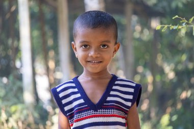 Kishoreganj, Bangladeş - 18 Aralık 2025 - Çizgili süveter giyen neşeli genç bir çocuk köy ortamında bir portre için gülümsüyor. Güney Asya 'daki bir çocuğun otantik yaşam tarzı tasviri.