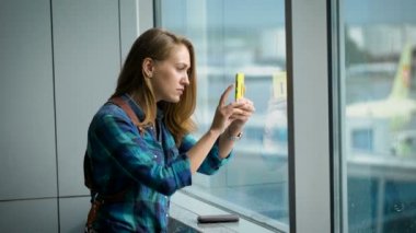 Çekici genç kadın alarak fotoğraf mobil cep telefonu Havaalanı Salonu büyük pencere oturmak ile portresi