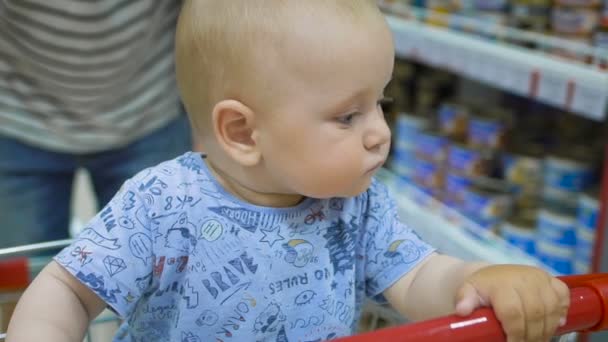 Pequeno bebê sentado em um carrinho de supermercado, enquanto seu pai escolhe compras . — Vídeo de Stock