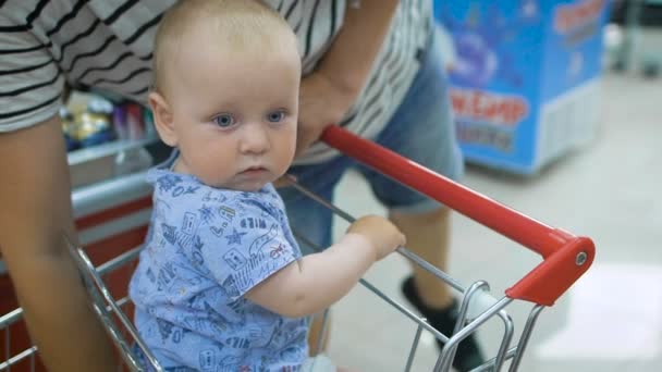 Малыш сидит в тележке с продуктами в супермаркете, в то время как его отец платит за покупки на кассе — стоковое видео