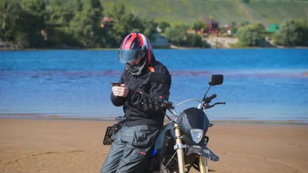 Motociclista sulla riva del fiume, vicino alla sua moto, sta cercando il telefono . — Video Stock