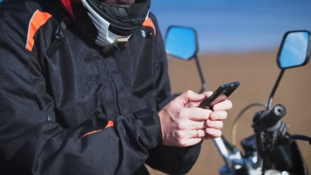 Motociclista en la orilla del río, junto a su bicicleta, que está buscando el teléfono . — Vídeos de Stock