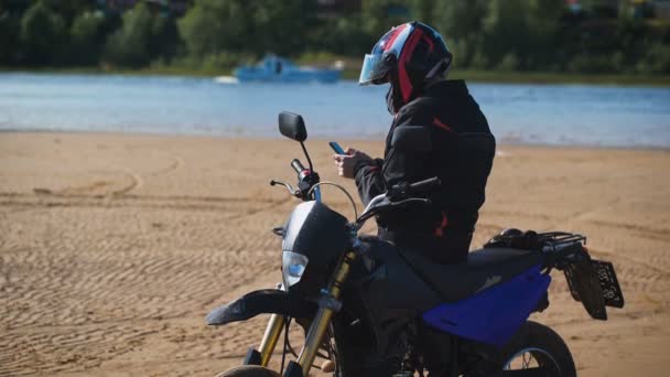 Motorkář na břehu řeky, vedle jeho kolo, dívá se po telefonu. — Stock video