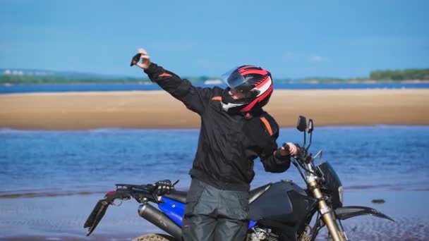Biker sur la rive de la rivière, à côté de sa moto, fait la photo au téléphone . — Video
