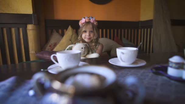 Seorang gadis kecil yang bahagia sedang duduk di sofa di kafe dan memeluk boneka kelincinya . — Stok Video