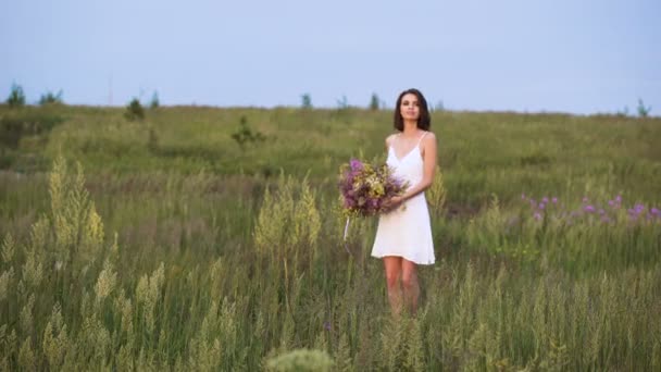 Young girl walks on a sunset on summer field with a bouquet of flowers. — ストック動画