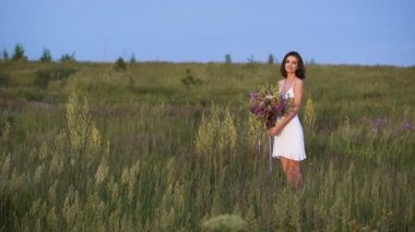 Young girl walks on a sunset on summer field with a bouquet of flowers.