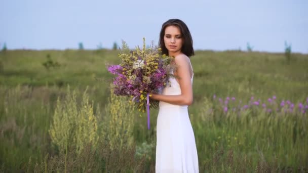 Jeune fille marche sur un coucher de soleil sur un champ d'été avec un bouquet de fleurs . — Video