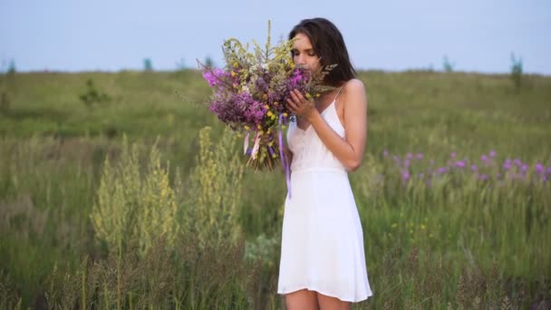 Young girl walks on a sunset on summer field with a bouquet of flowers. — Stok video