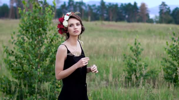 Menina bonita no vestido curto preto com a coroa de flores na cabeça no campo verde . — Vídeo de Stock