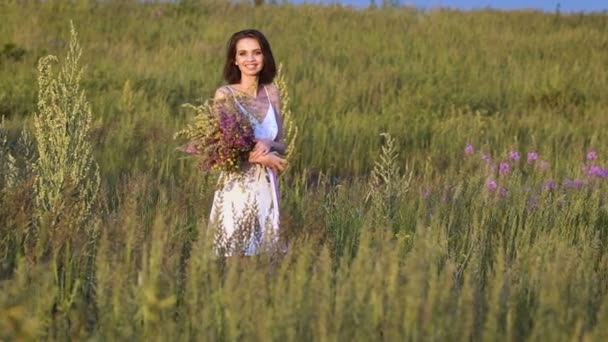 Une jeune femme marche sur un champ vert avec un bouquet de fleurs et souriant — Video