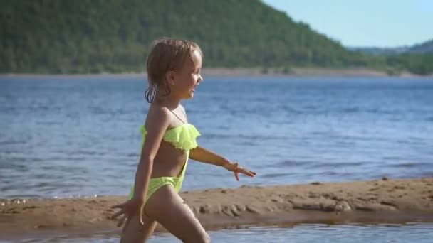 Χαριτωμένο μικρό ξανθό κορίτσι που τρέχει από μια λίμνη με τα πράσινα βουνά στο φόντο — Αρχείο Βίντεο