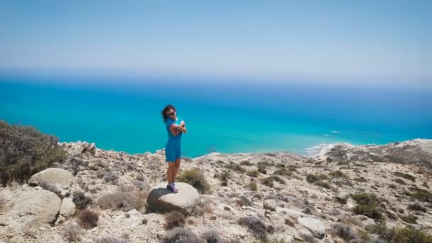 Una mujer joven en vestido azul de pie en la montaña con la hermosa vista al mar — Vídeo de stock