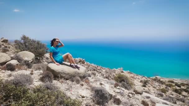 Μια νεαρή γυναίκα στο μπλε φόρεμα κάθεται στο βουνό με την όμορφη θέα στη θάλασσα — Αρχείο Βίντεο