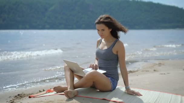 Uma bela sensual jovem senhora está sentada e sorrindo em uma praia usando laptop — Vídeo de Stock