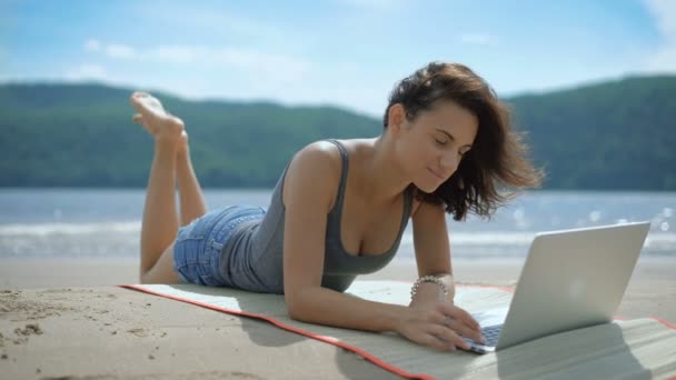 Μια όμορφη σέξι νεαρή κοπέλα ευτυχισμένος είναι laying επί της tommy σε μια παραλία που χρησιμοποιούν φορητό υπολογιστή — Αρχείο Βίντεο