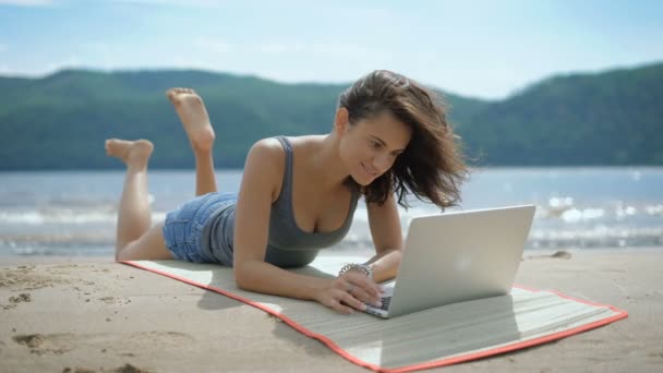 Μια όμορφη σέξι νεαρή κοπέλα χαμογελαστή είναι laying επί της tommy σε μια παραλία που χρησιμοποιούν φορητό υπολογιστή — Αρχείο Βίντεο