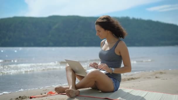 Een mooie sexy jonge inspirated dame zit bij een strand met laptop — Stockvideo