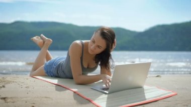 Güzel seksi genç düşünceli Bayan ona tommy dizüstü bilgisayar kullanarak bir kumsalda yatıyor