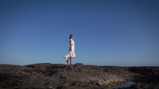 Mujer joven delgada en un vestido griego blanco camina en una orilla rocosa. — Vídeos de Stock
