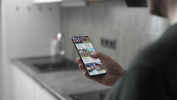 Un hombre elige un nuevo hogar para sí mismo a través de una aplicación en un teléfono inteligente. Selección remota y compra de bienes raíces desde el teléfono. — Vídeos de Stock