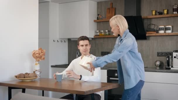 Hermosa rubia le da a su novio un regalo de cumpleaños. Jóvenes alegres en casa en la cocina. — Vídeo de stock