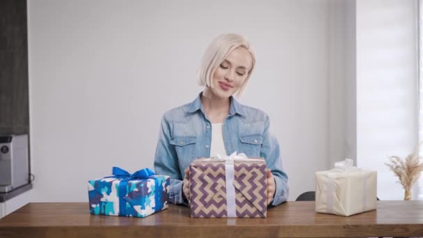 Όμορφη γυναίκα στο σπίτι στο τραπέζι εξετάζει ένα δώρο. Ένα κουτί σε κουτί δώρου στα χέρια μιας ξανθιάς.. — Αρχείο Βίντεο