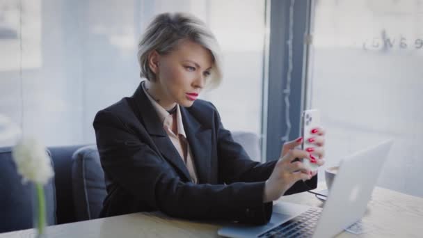 O blondă într-o cafenea cu un telefon. O femeie de afaceri face un selfie . — Videoclip de stoc