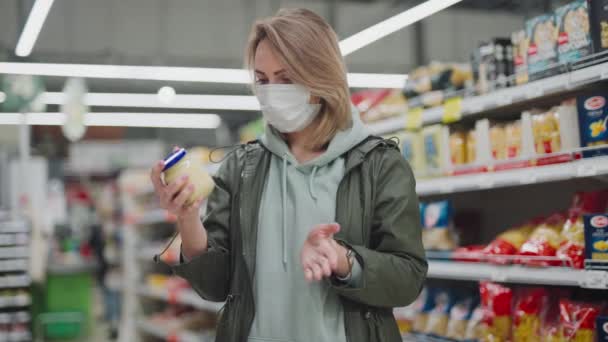 Frau mit Schutzmaske im Supermarkt. Eine Hausfrau im Regenmantel sucht sich Essen aus. — Stockvideo