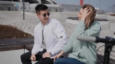 Bir adam ve bir kadın bankta oturmuş sohbet ediyorlar. Çiftin yanında elektrikli scooterları var..