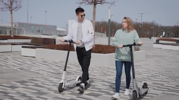 Genç bir adam ve kadın sokakta iletişim kuruyor. Çift gezintiye giderken elektrikli scooterları da yanlarında götürdü.. — Stok video