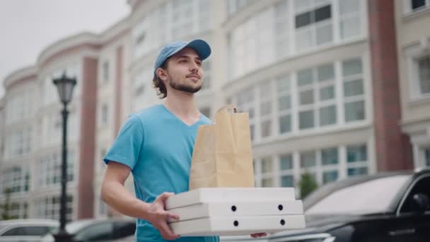 Mladý poslíček jídla prochází moderní, příjemnou čtvrtí. Muž v čepici a tričku doručuje pizzu a potraviny — Stock video